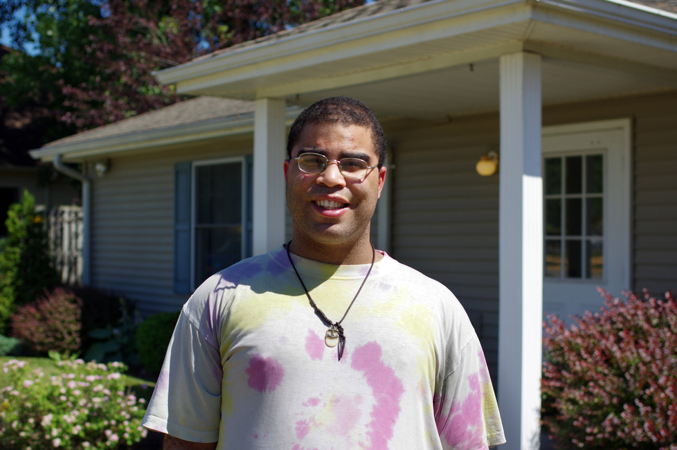 Young man wearing glasses and a tie dye shirt standing in front of house on a sunny day.